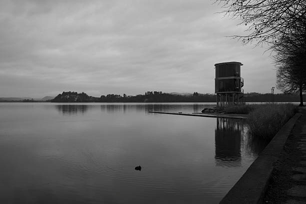 Black and white image of a minimalist lakeside pier leading toward calm water, with a modern structure on the shore. The quiet scene, soft tones, and open space convey stillness, solitude, and balance, creating a strong sense of calm and contemplation.