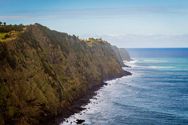 Scenic view of the rugged coastline of Nordeste on Sao Miguel Island, Portugal. The image features steep green slopes dropping into the Atlantic Ocean under a moody sky.