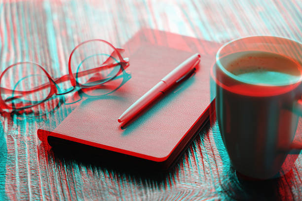 A creative conceptual image featuring a work desk setup with a leather notebook pen glasses and a mug of coffee rendered with a vibrant cyan and red double exposure effect on a wooden texture