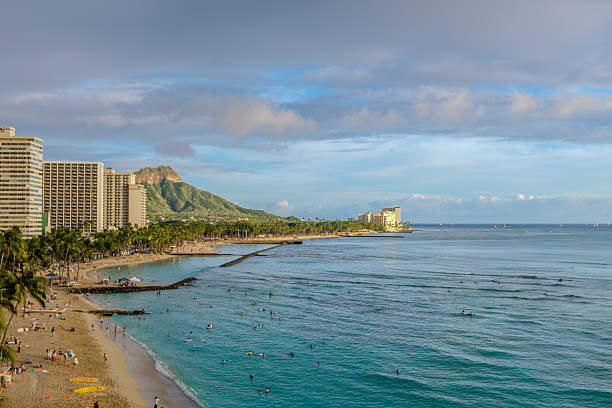 Waikiki shoreline at golden hour with Diamond Head in the distance, gentle surf and swimmers filling the turqoise water beneath a pastel sky.