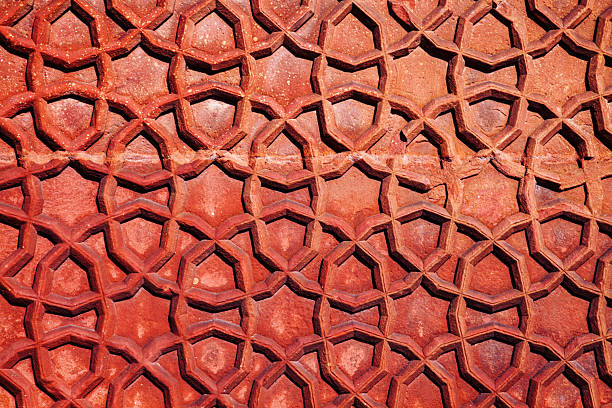This detailed close-up highlights the masterful craftsmanship of geometric star carvings on a red sandstone wall at the historic Red Fort in Agra, India. The interplay of bright sunlight and deep shadows emphasizes the intricate textures and architectural grandeur of Mughal design.