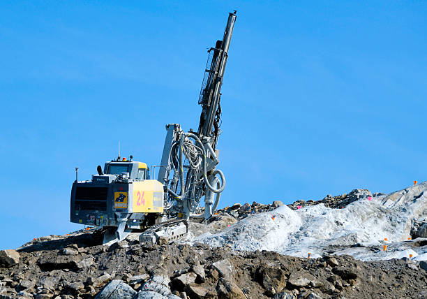 Nuuk / Godthåb, Sermersooq, Greenland: profile view of an Epiroc SmartROC T40 surface drill rig operating on a rugged site in the capital of Greenland. The heavy machinery, branded with the logo of the Danish contractor Munck, is positioned on uneven, grey bedrock against a clear, cloudless blue sky. Small orange survey markers are visible on the ground, outlining the drilling pattern for upcoming blasting work. This hydraulic top-hammer rig is a staple in Arctic civil engineering projects, facilitating infrastructure development in the challenging geological and meteorological environment of Nuuk.