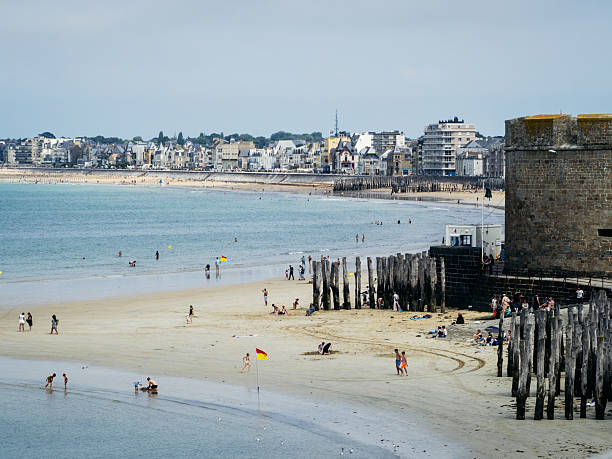 People enjoy a summer day on the main beach of Saint-Malo, Brittany, with historic seawalls, wooden breakwaters, and the town skyline visible in the background.