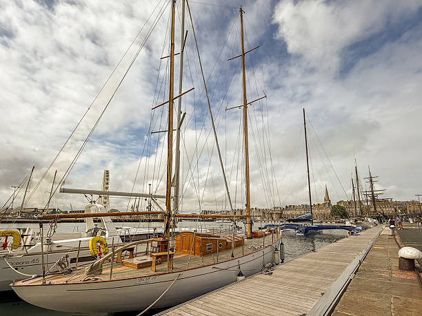 Traditional sailboats are moored along a wooden pontoon  in the harbor of Saint-Malo, Brittany, with historic city walls and waterfront buildings visible in the distance.