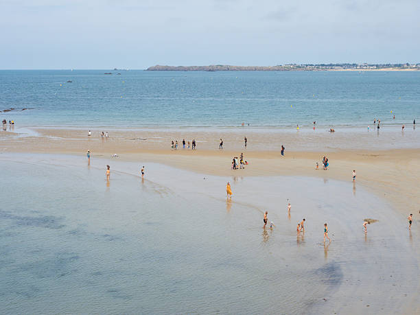 People walk and wade across shallow water at low tide on a wide sandy beach in Saint-Malo, Brittany, enjoying a calm summer day along the French coast.