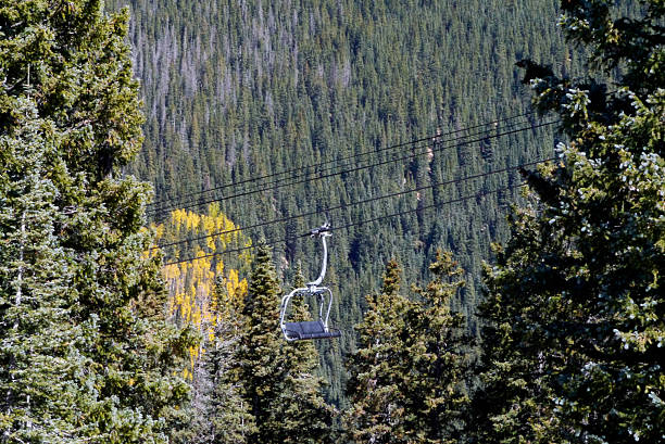 During the off-season, Aspen Peak is available for hiking and seeing the fall leaves turning. Here, an empty chair on the ski lift passes through an evergreen grove. Santa Fe, NM 2025