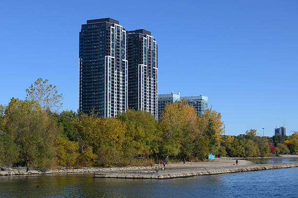 Tall residential skyscrapers stand against a blue sky, overlooking a tranquil waterfront with a concrete pier and vibrant autumn trees in Etobicoke, Toronto