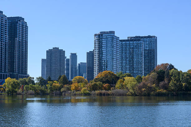 Modern residential buildings in Etobicoke, Toronto, standing tall behind vibrant autumn trees bordering a calm lake under a clear blue sky, illustrating urban natural beauty