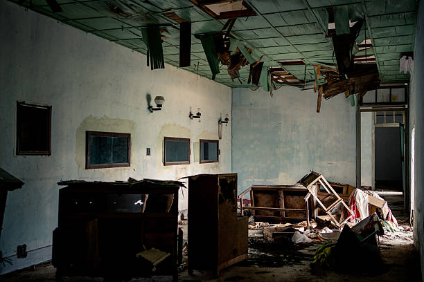 Striking photograph of a severely damaged abandoned room featuring a partially collapsed ceiling with hanging debris, exposed beams, broken plaster, and scattered overturned desks, chairs, and wreckage on the floor under dim, diffused light. This evocative image symbolizes structural failure, the passage of time, and the haunting beauty of decay, ideal for horror films, dystopian art, historical documentation, or themes of urban abandonment and impermanence. High-resolution details showcase intricate textures of destruction, peeling paint, and chaotic composition for maximum visual impact in editorial, cinematic, or fine art contexts.