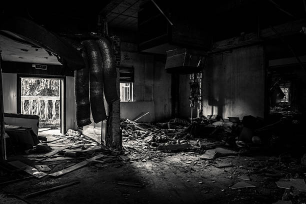 High-contrast black and white photograph of a heavily damaged abandoned interior featuring collapsed sections, hanging flexible ducts, overturned furniture, piles of rubble, broken plaster, and intense shafts of light streaming through shattered windows and doorways, creating stark shadows and a powerful sense of chaos and abandonment. This dramatic image powerfully conveys destruction, the aftermath of neglect or disaster, and the haunting aesthetic of urban ruins, perfect for horror, dystopian art, thriller visuals, documentary ruin series, fine art prints, or editorial features on impermanence and decay. High-resolution monochrome with extreme tonal range, intricate textures of debris and destruction, and cinematic lighting for maximum emotional impact and visual storytelling.