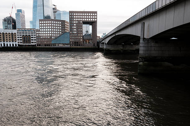 London bridge extending over the river thames with the modern city skyline featuring the shard, reflecting urban architecture and transport infrastructure under a cloudy sky