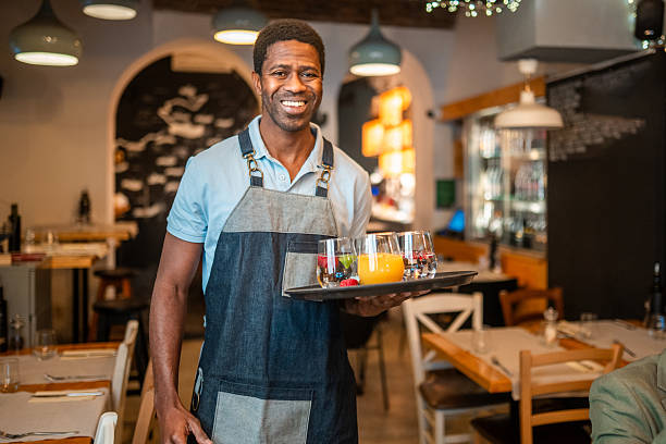 Male waiter in a cozy brunch bistro holding a tray full of drinks. He is smiling as he stands in the dining area, ready to serve customers. The bistro features a warm, inviting atmosphere with wooden tables and ambient lighting.