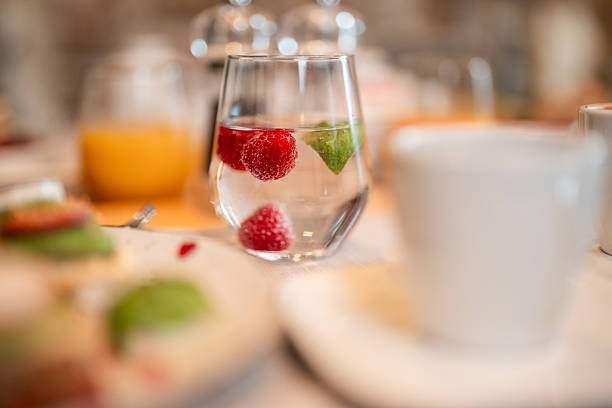 Clear glass of water with red raspberries and fresh green mint leaves, served on a white tablecloth at a cozy bistro brunch table. Other blurred brunch items, including orange juice and a plate with food, appearing in the background. A clean white teacup and saucer also visible nearby, suggesting a relaxed morning meal.