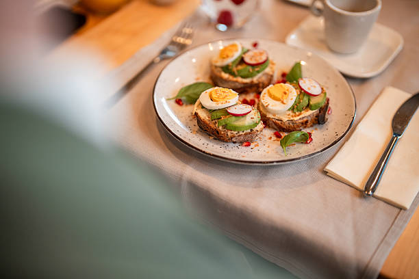 A healthy brunch meal set on a bistro table. A plate holds three avocado toasts, topped with boiled eggs, radish slices, and pomegranate seeds. Nearby, a coffee cup, cutlery, and a refreshing glass of water are arranged on the light-colored tablecloth.