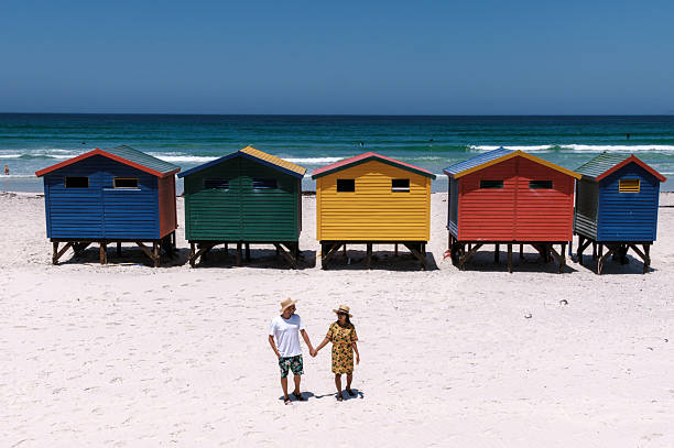 Enjoy the stunning Muizenberg beach in Cape Town, where visitors stroll hand in hand along the sandy shore. Brightly colored beach huts create a picturesque backdrop against the beautiful ocean.
