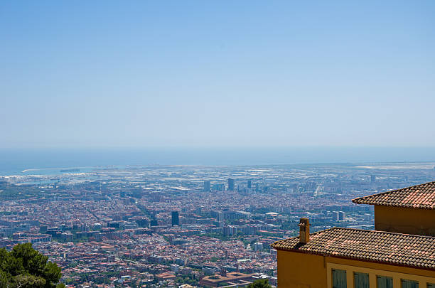 Wide aerial shot of a densely built coastal city with terracotta rooftops, clear blue sky, and distant sea horizon.