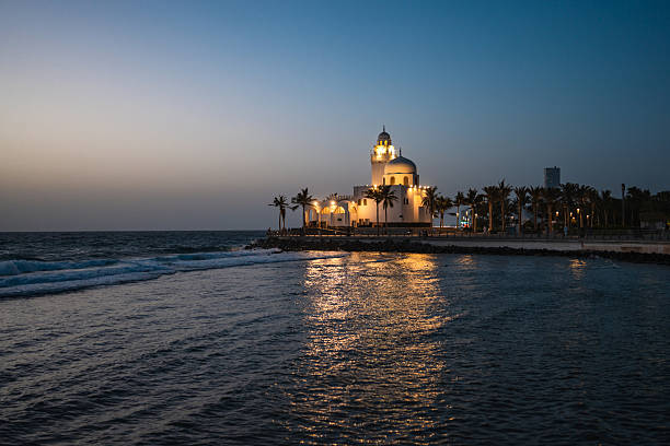 Al-Rahma Mosque, also known as the Floating Mosque, glowing softly at dusk on the Jeddah Corniche featuring a serene coastal view with gentle waves lapping against the shore under the evening sky.
