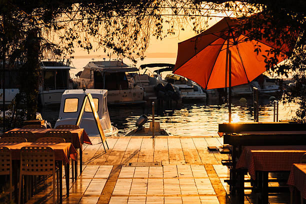 An inviting view of a wooden restaurant terrace with red checkered tablecloths located directly on the harbor. An orange parasol and silhouettes of boats in the background complete this warm summer evening scene.