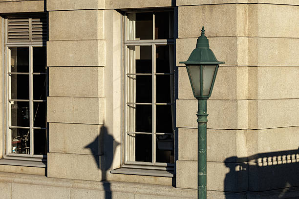 Vintage green streetlamp with European flair basks in low angle late afternoon sunlight, casting elongated shadows on to neoclassic stonework face of adjacent building.