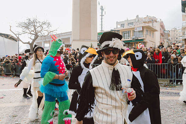 Xanthi, Greece - February 18, 2018: A group of adult carnival participants wearing humorous and colorful costumes walk along a cobblestone street during the annual Xanthi Carnival parade, including men dressed as penguins, a green fantasy character, and a man in a vintage-style outfit with a top hat, while large crowds of spectators watch behind metal barriers in the city center