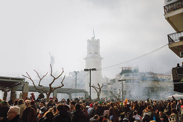 Xanthi, Greece - February 18, 2018: Large crowd gathers in the central square of Xanthi during the annual carnival celebrations as thick smoke drifts through the city creating a dramatic atmosphere around the iconic clock tower while thousands of people in costumes celebrate and watch the festivities