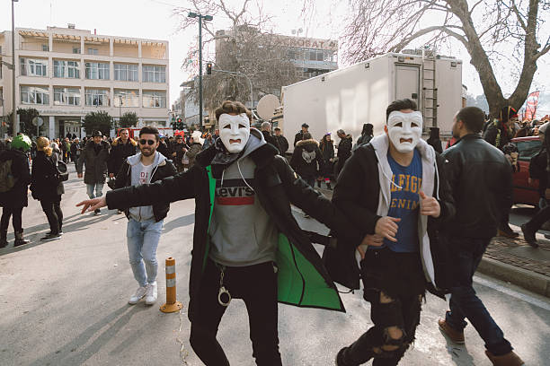 Xanthi, Greece - February 18, 2018: Young men wearing white theatrical masks run and gesture playfully in the streets of Xanthi during the annual carnival celebrations as crowds of people gather in the background creating a lively and chaotic atmosphere typical of the famous Greek carnival event