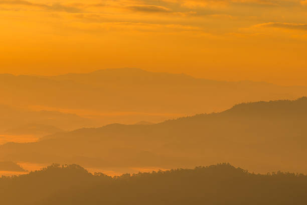 Layered mountain ridges emerge through thick mist during a vibrant orange sunrise. This minimalist landscape offers vast copy space, perfect for nature, travel and tranquility background themes.