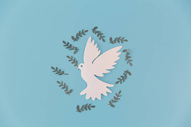 White paper cut dove surrounded by an olive leaf wreath on a blue background. A minimalist symbol of global peace, unity and hope with ample copy space for humanitarian or spiritual projects.