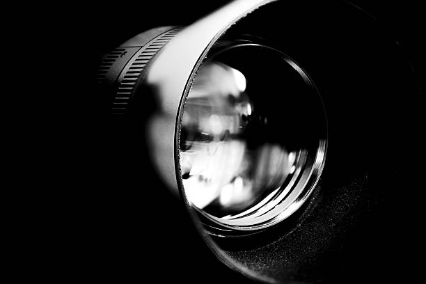Close-up view of a camera lens in black and white, emphasising glass elements, reflections, and precision engineering against a dark background.