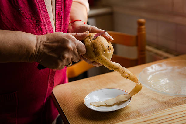 Close-up of an adult woman’s hands peeling a raw potato with a knife over a small plate on a wooden kitchen table. Potato skins are visible, showing a traditional home cooking preparation process.