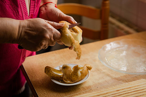 Close-up of an adult woman’s hands peeling a raw potato with a knife over a small plate on a wooden kitchen table. Potato skins are visible, showing a traditional home cooking preparation process.