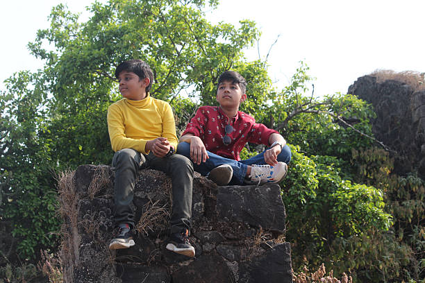Two young Indian boys sitting on a low wall in a park, enjoying the tranquility of their surroundings while sharing moments of laughter and friendship amidst nature's beauty