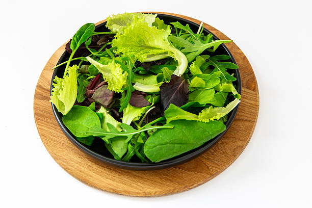 A bowl filled with various types of green leafy vegetables is placed on a round wooden board. The setting suggests a fresh salad ready for a meal. This scene is simple and direct.