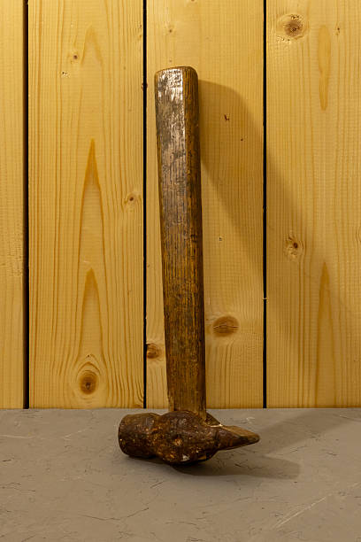 Old hammer with wooden handle stands upright on concrete surface. Vertical wooden planks visible behind tool.