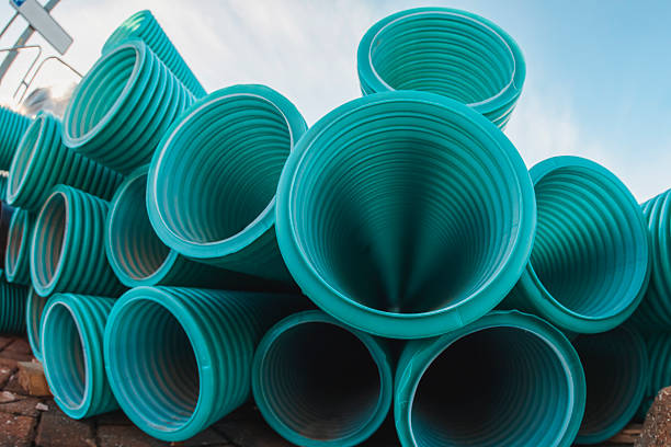 Teal pipes are arranged in a stack at a construction site under a clear sky. The sunlight reveals the texture and pattern of the pipes. Construction materials are visible.