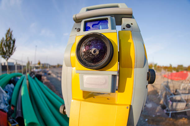 A surveying instrument stands on a construction site. Green pipes are stacked in the background. The sky is clear, and the sun is shining down on the area.