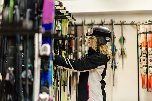 A beautiful young woman dressed in ski wear is choosing skis in a ski room.