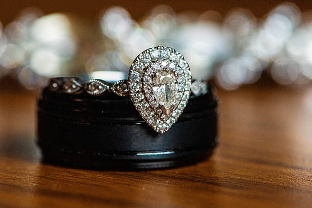 Elegant pear-cut diamond engagement ring positioned on top of masculine black titanium wedding band against sparkling bokeh background. Shows bride and groom's rings together in symbolic union. Represents partnership unity, feminine and masculine joining, contrast creating harmony, two lives becoming one, eternal commitment, complementary strengths, and the beautiful balance of differences. Symbolizes marriage covenant, inseparable bond, layered love, stacking of lives together, and the sparkle of shared future. Metaphor for compatibility through contrast and unity in diversity. Perfect for illustrating marriage commitment, wedding symbols, partnership unity, eternal love, complementary relationships, wedding planning, and covenant bonds.