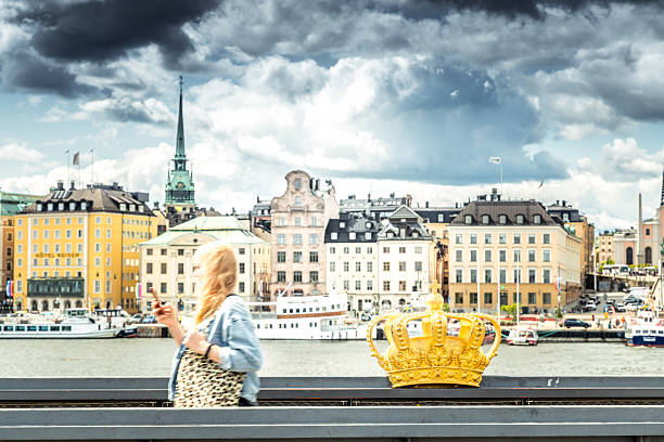Stockholm, Sweden - June, 19, 2025:   A woman walks on the Skeppsholmsbron with a smartphone in her hand with Gamla Stan in the background. Cloudy unpredictable weather