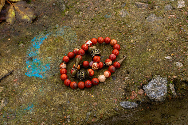 Beaded bracelet with red stones and metal elements rests on rough concrete. Leaf and small stones visible nearby.