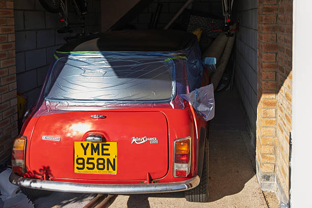 Huntingdon, UK: Mini Cooper Innocenti 1300 Export having a partial resprayA classic red Mini Cooper automobile is parked inside a dimly lit home garage, viewed from the rear to highlight the compact design,  taillights, and yellow UK license plate reading YME 958N. The trunk area appears slightly ajar with reflective surfaces catching light, surrounded by storage shelves stocked with boxes, papers, and household items indicating everyday use. No people are present. The scene conveys a sense of nostalgic automotive storage in an urban residential setting.