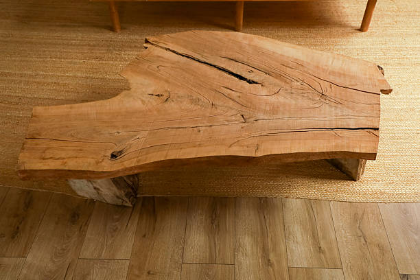 A large wooden table sits in a room with a soft rug underneath. The table has natural features and markings. Sunlight fills the space showing the grain of the wood.
