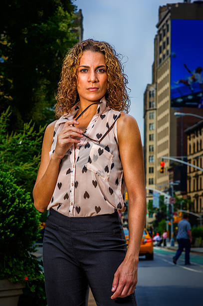 Woman Standing Confidently in Cityscape. A woman with curly hair stands confidently in an urban environment. She is dressed in a stylish sleeveless blouse and trousers, surrounded by busy city life.