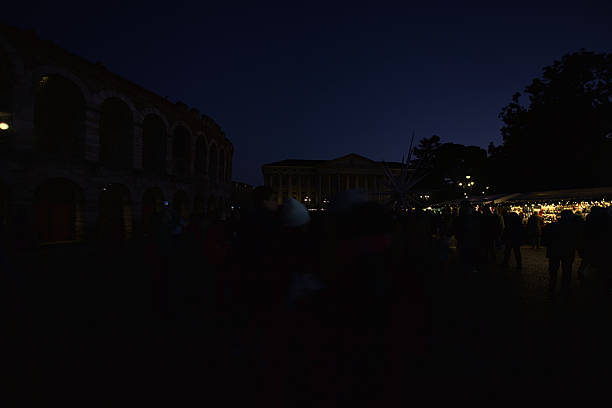 Verona, Italy - 8TH DECEMBER 2025 - Lit Christmas market stalls next to the Arena di Verona and the town hall in Bra square at night, with people passing by, strolling, and browsing stalls