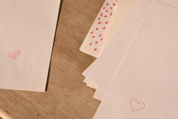 Horizontal photo of white paper sheets on a wooden table. On the left, a sheet with a small heart drawn and slightly filled with pen. On the right, a stack of papers with a heart outline and small painted hearts on lower sheets.