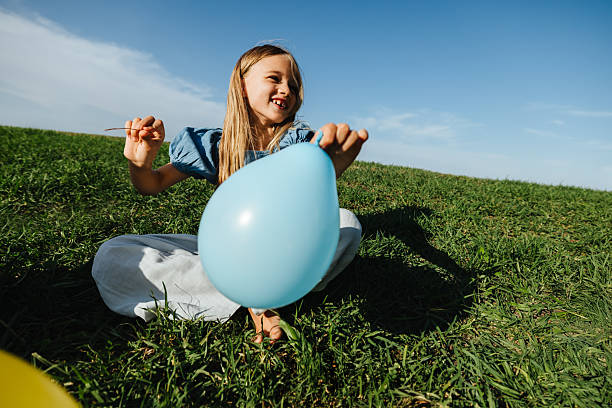 A girl sits on grass outdoors, holding a blue balloon and string, smiling as she plays in an open field.