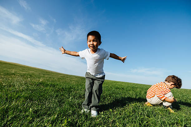 A young boy runs with arms outstretched across a green field while another child sits nearby, exploring the grass under a clear blue sky.