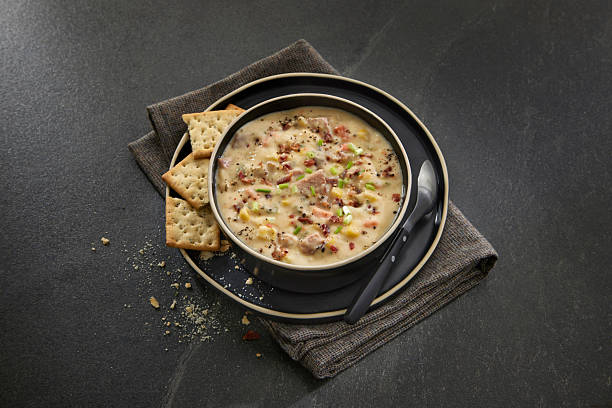 Creamy ham and corn chowder garnished with green onions and black pepper, served in a bowl with crackers on a dark background.