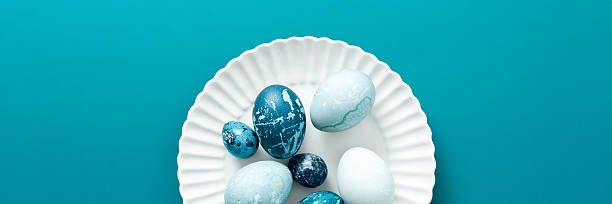 Colorful blue Easter eggs are artistically arranged on a white plate resting on a vibrant teal surface, capturing the essence of spring festivities and holiday traditions, banner