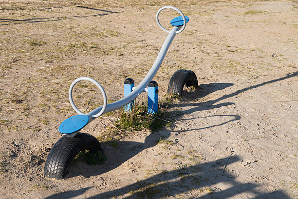 A brightly lit blue and white seesaw sits empty on a dry, sandy playground. The simple metal teeter-totter, with its round handles and plastic seats, rests on old tires embedded in the ground, casting long shadows under the clear daylight. Sparse green and dry grass peeks through the dusty earth, suggesting an unused or perhaps a vintage play area. It evokes feelings of childhood memories, solitude, and simpler times.